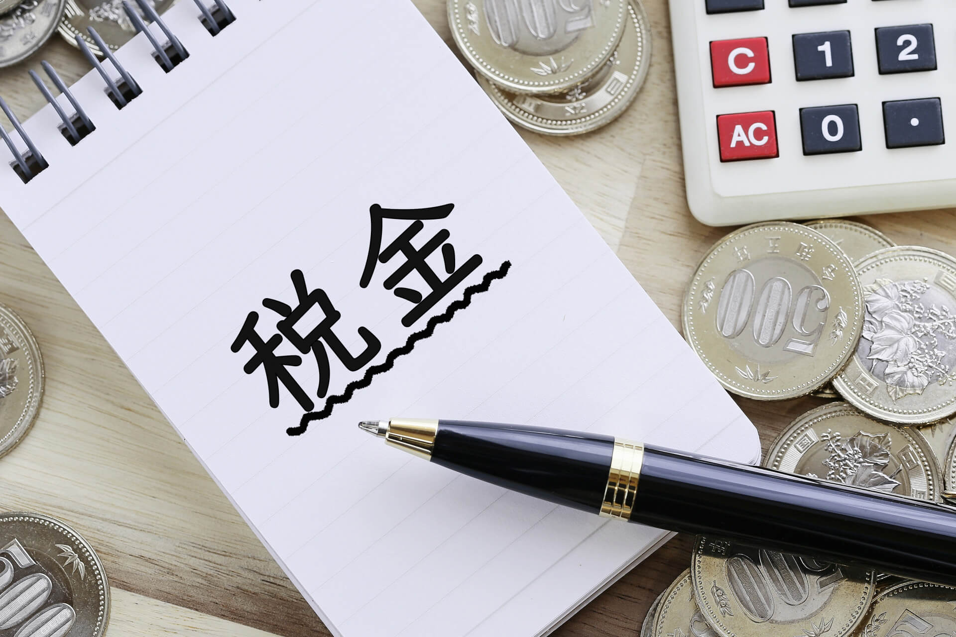 「税金」と書かれたノートと硬貨、電卓｜空き家の固定資産税を表すイメージ